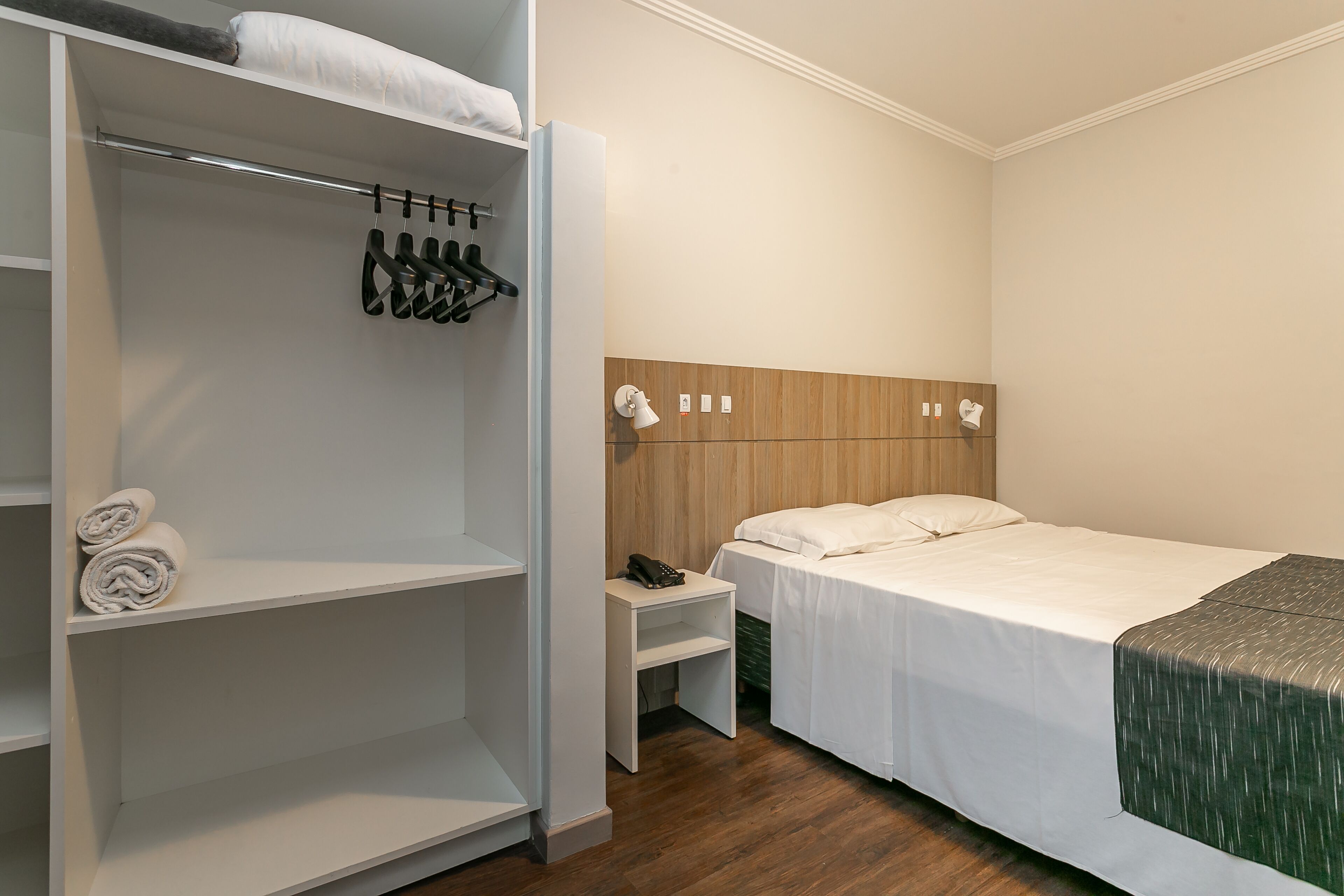 Habitación de hotel con una cama bien hecha, una mesita de noche con lámpara, cabecero de madera y armario con zapatos colgados.