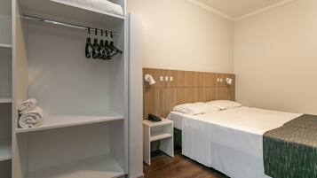Habitación de hotel con una cama bien hecha, una mesita de noche con lámpara, cabecero de madera y armario con zapatos colgados.