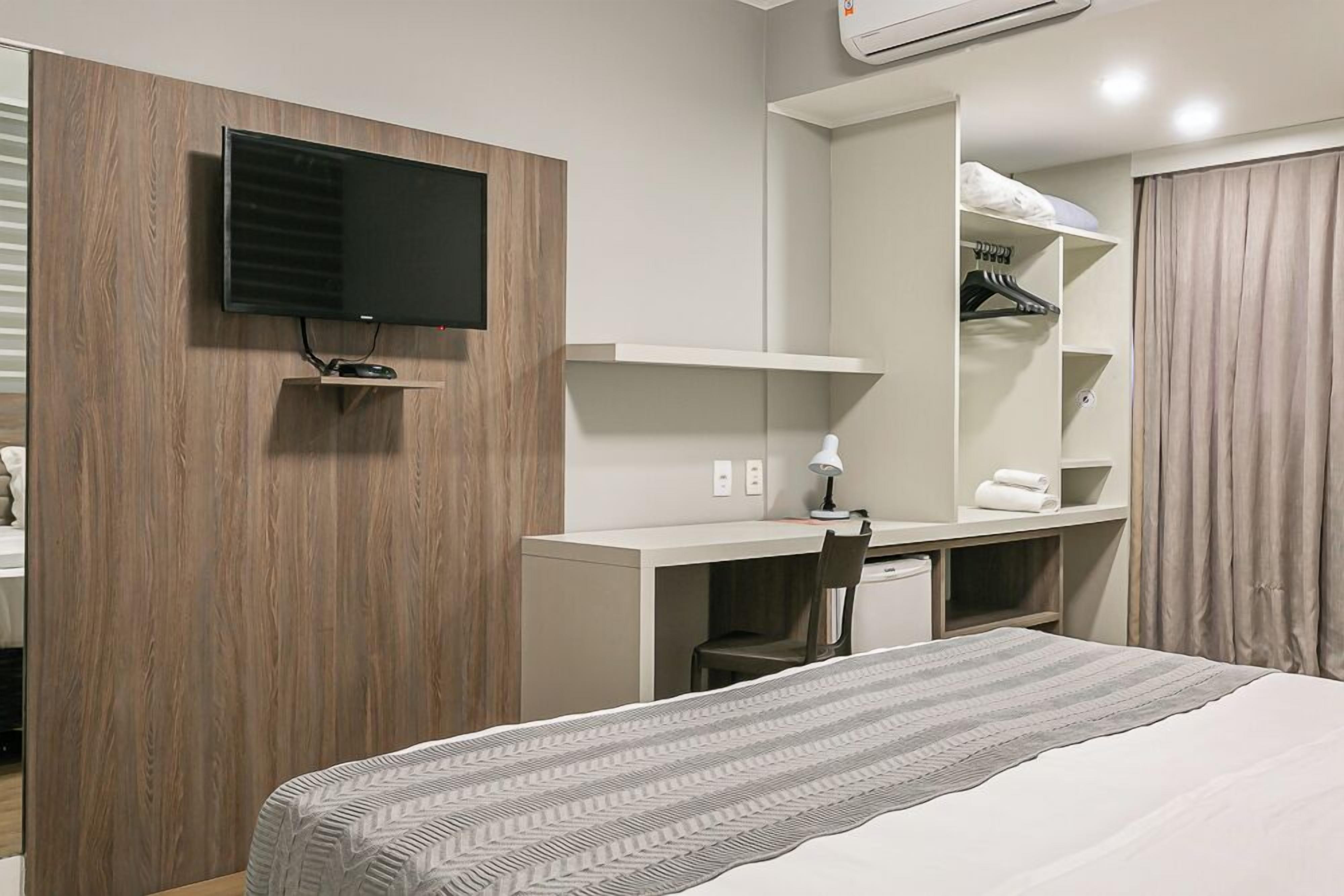 A hotel room with a bed, a desk with a chair, a television, a wardrobe, and a mirror.