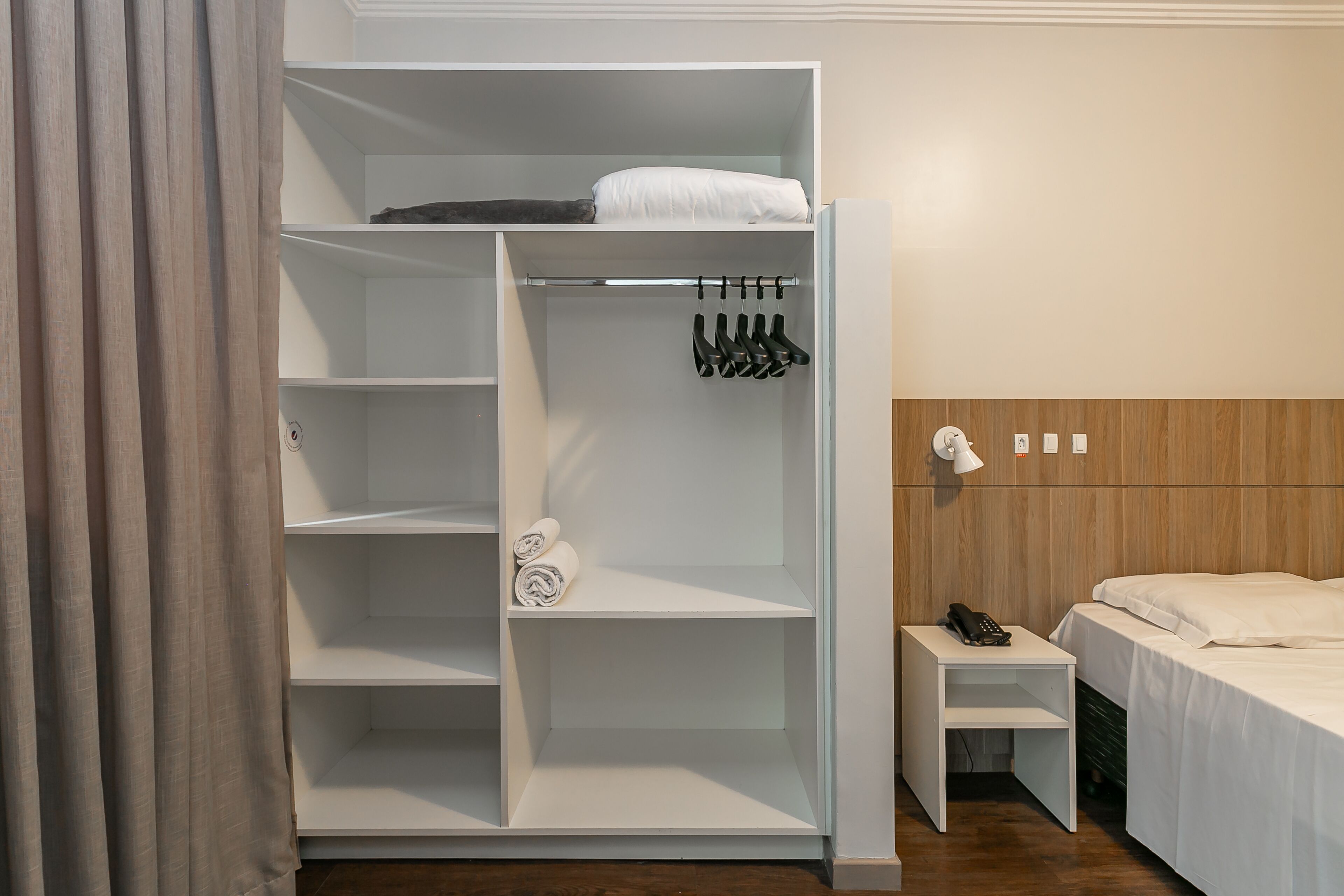 Habitación de hotel con un armario blanco, una cama, una mesita de noche con un teléfono y un cabecero de madera.