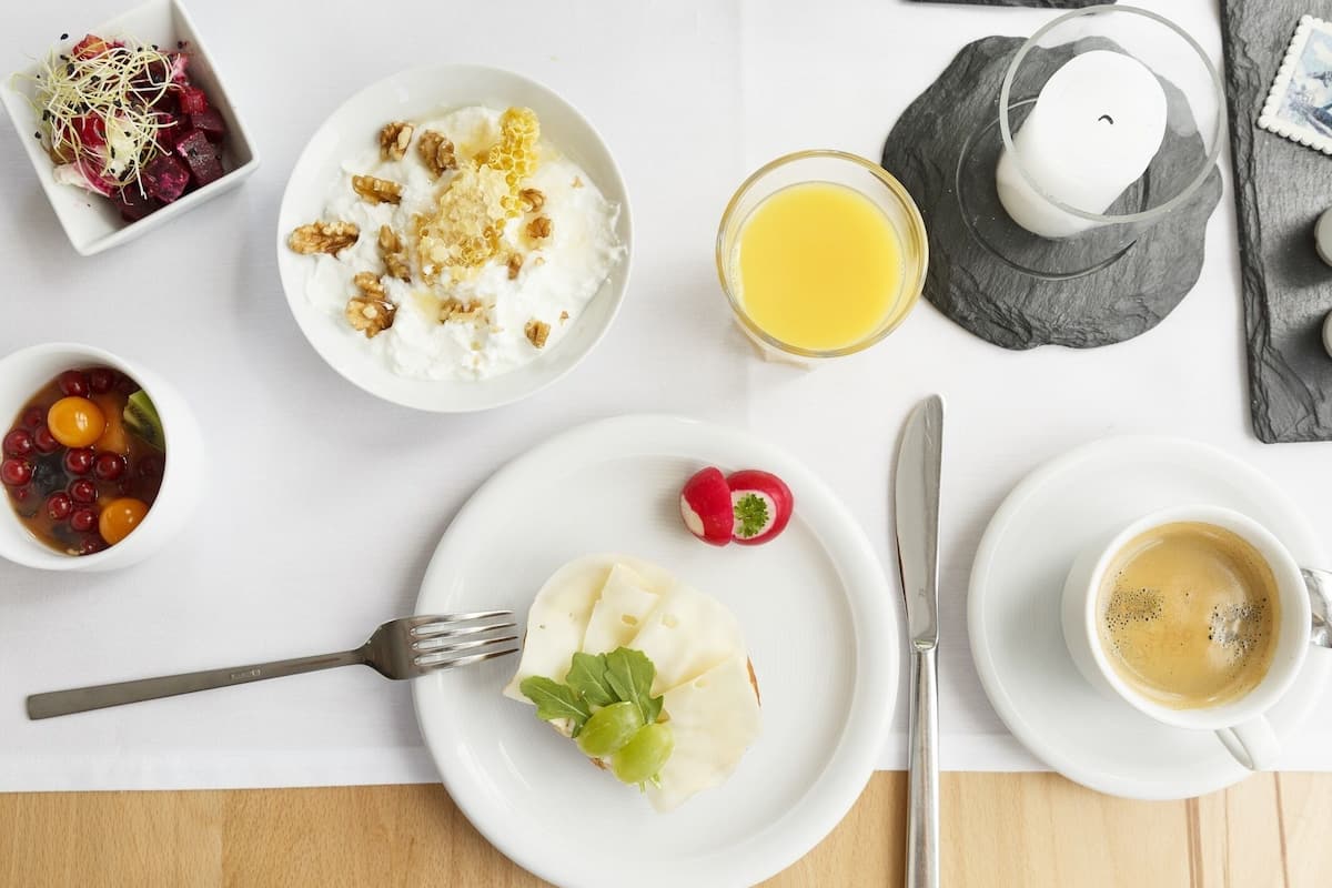 Un desayuno con yogur cubierto de nueces y miel, un vaso de jugo de naranja, una taza de café y un plato con un rábano y una ensalada pequeña.