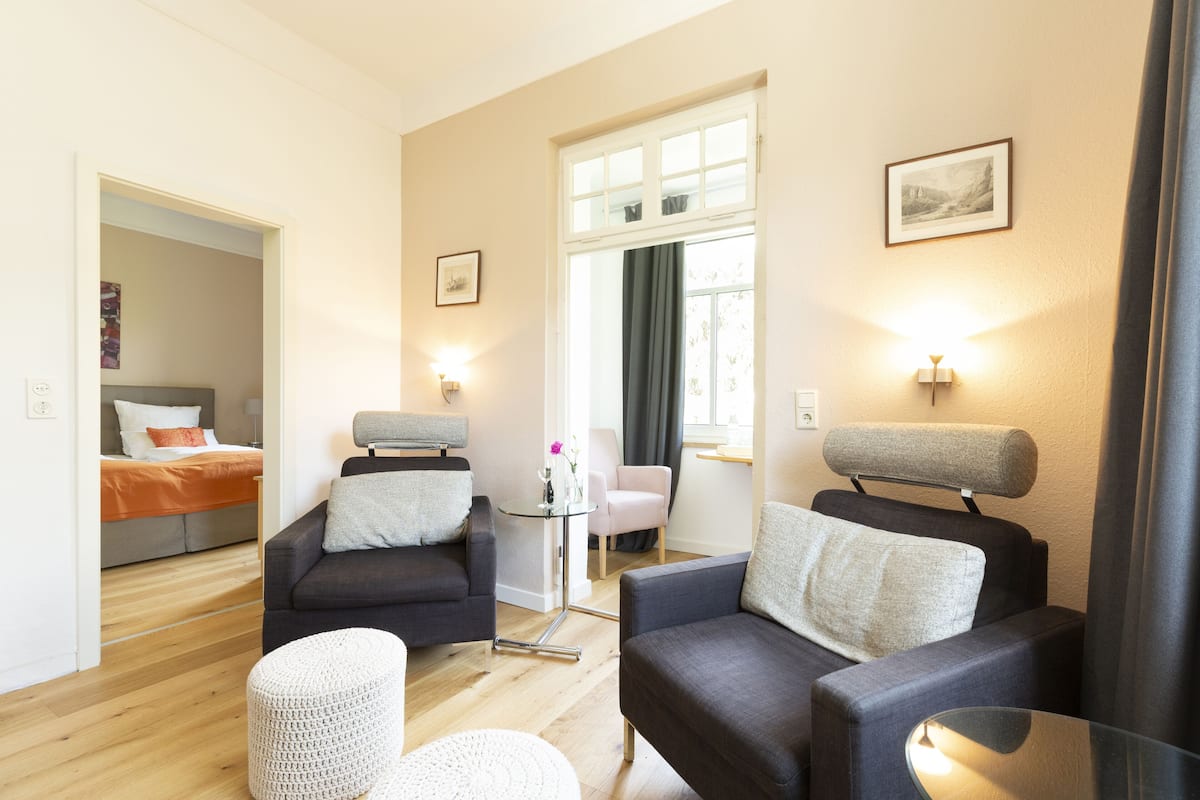 Una sala de estar moderna con un sofá, una mesita redonda y una pequeña mesa auxiliar. Se ve una cama con un edredón naranja al fondo.