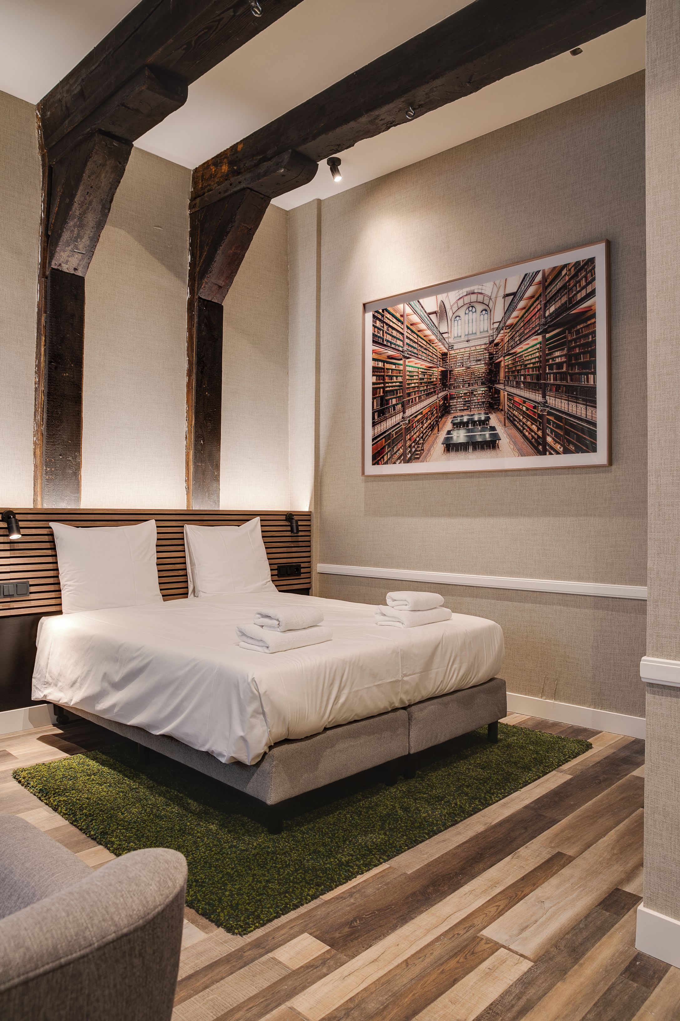 Een moderne slaapkamer met een groot bed, een hoofdbord met horizontale lijnen, een vloerkleed van kunstgras en een gekaderd beeld van een bibliotheek aan de muur.