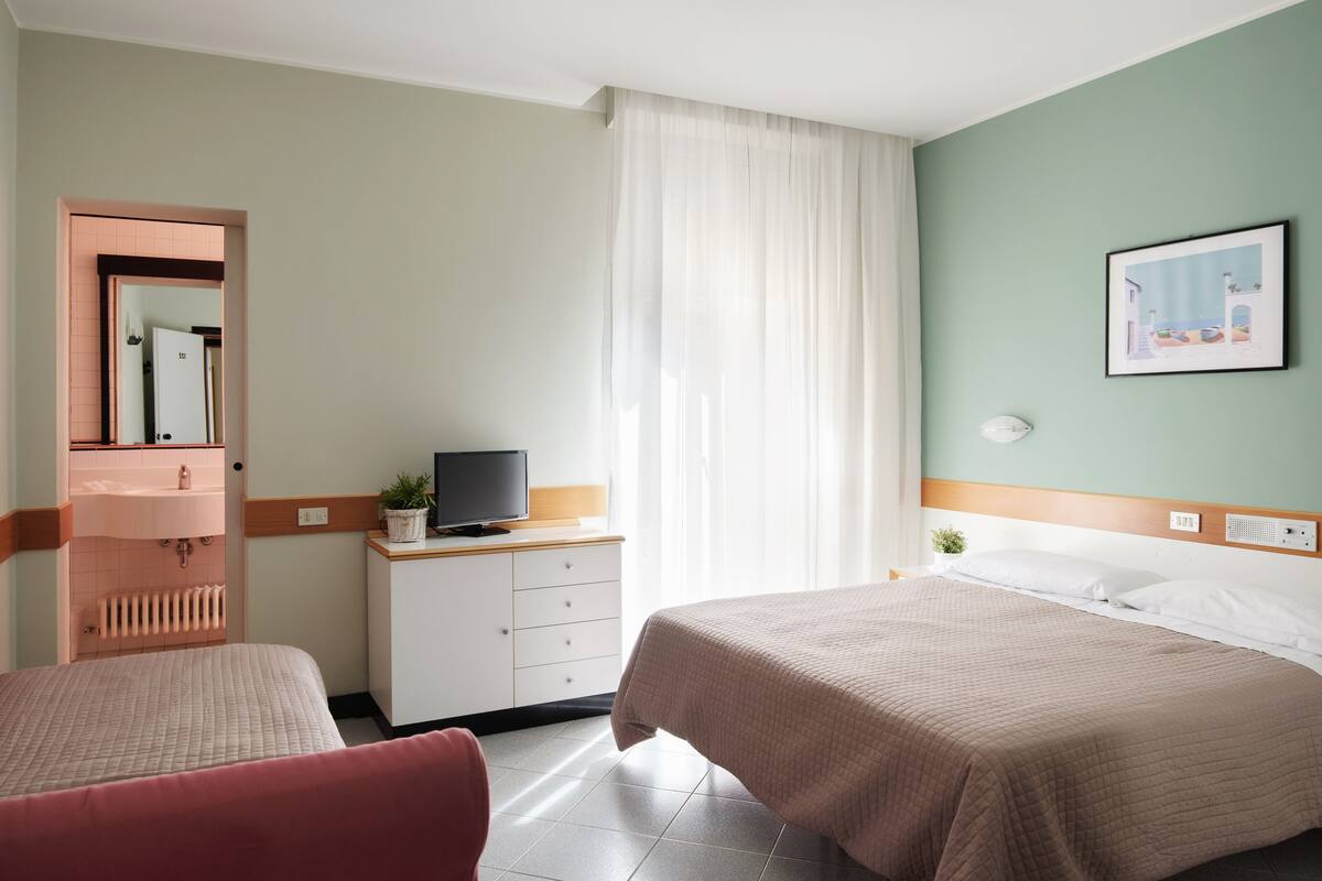 A hotel room with two beds, a television on a nightstand, a bathroom with a pink sink, and a window with sheer curtains.