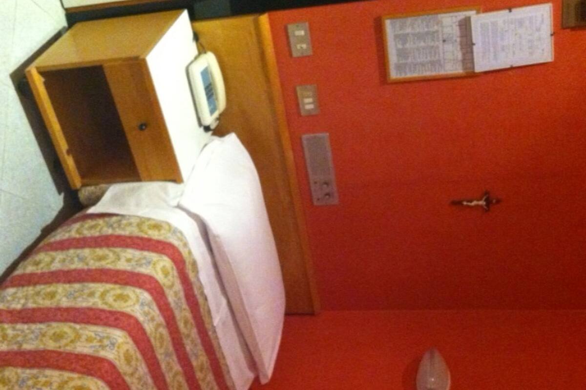 A hotel room with a bed, a bedside table, a telephone, and a wall-mounted information board.