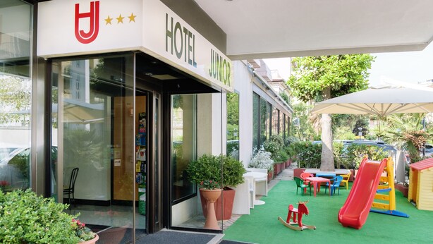 A hotel entrance with a glass door, a sign reading "HOTEL JUNIOR", and a children's play area with a slide and climbing structure.