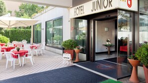 A hotel entrance with a sign reading "HOTEL JUNIOR" and a "Come in" sign. There are outdoor tables and chairs, and potted plants near the entrance.