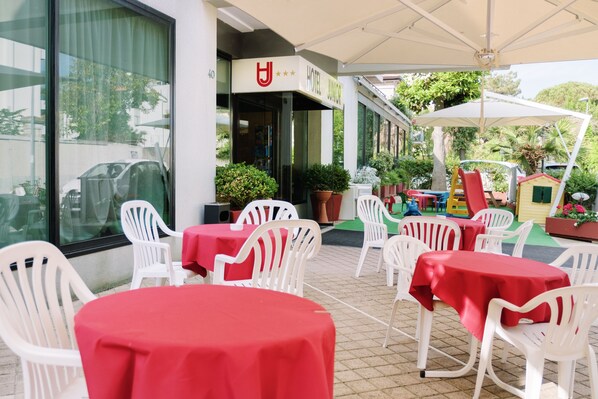 Un área de patio al aire libre con sillas y mesas de plástico blanco, un espacio de juegos para niños y un letrero de hotel.