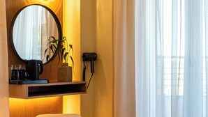 A bedroom with a round mirror, a small table with a plant, a hairdryer, and a window with sheer curtains. - Zante Pantheon Hotel (Zakynthos)