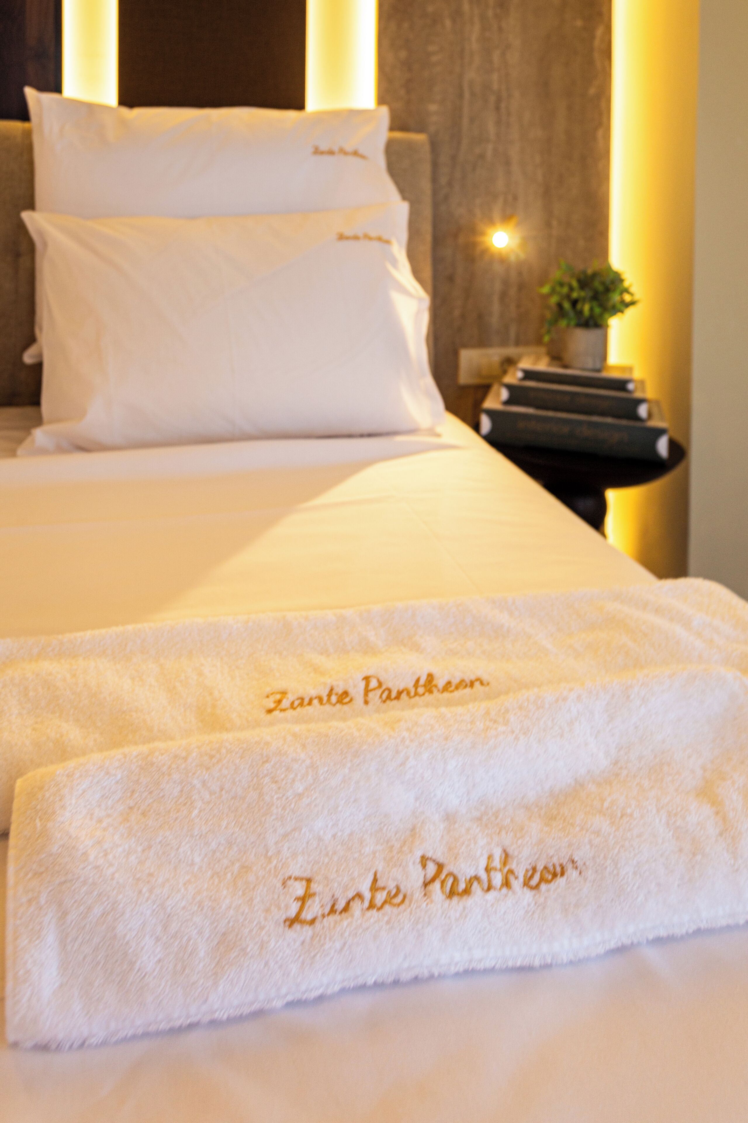 A neatly made bed with white linens and two towels with 'Zante Pancreon' embroidered on them.
