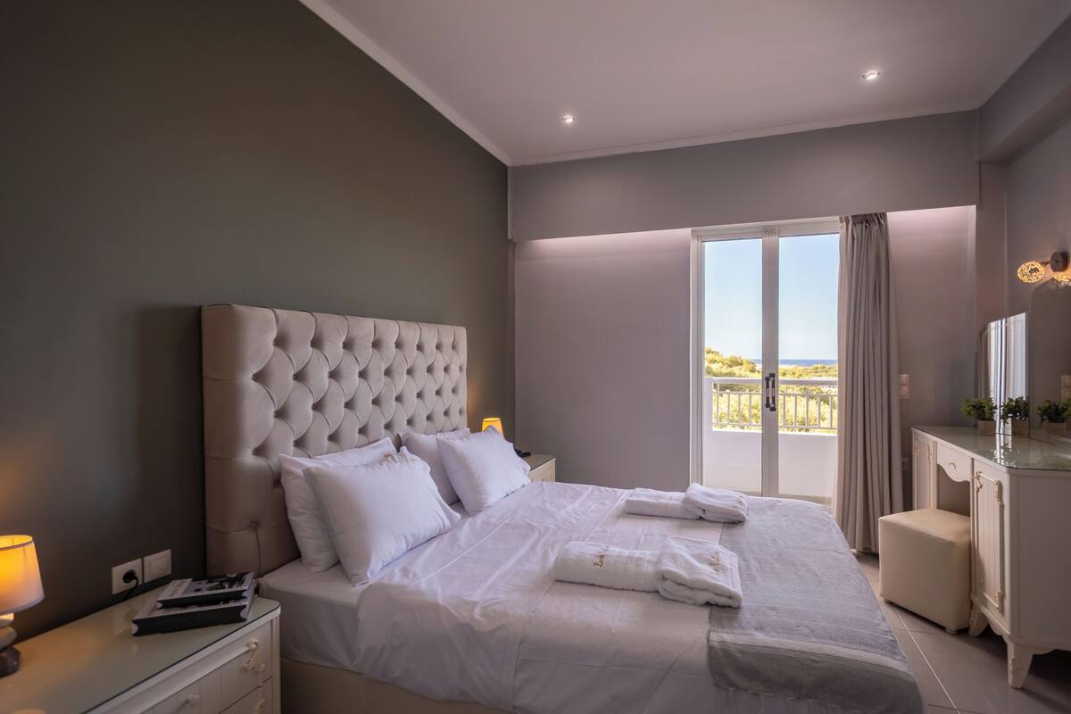 A bedroom with a tufted headboard, a large bed with white linens, a bedside table with a lamp, and a view of the outdoors through a sliding door.