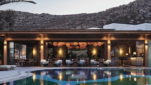 A poolside area with seating and a bar, surrounded by a mountainous landscape. - Myconian Sunrise Relais & Chateaux Hotel (Mykonos)