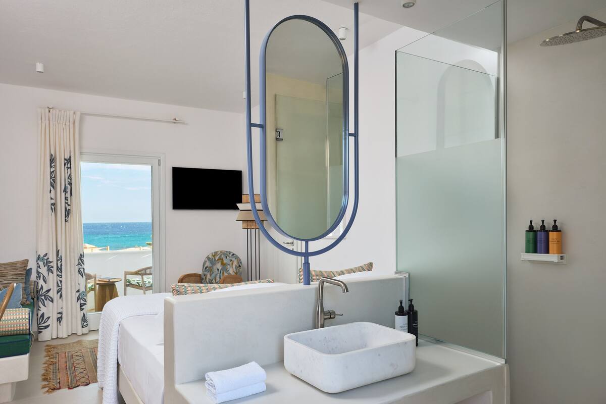 A modern bathroom with a large mirror, a white sink, and a shower. There is a TV mounted on the wall and a view of the ocean through the window.