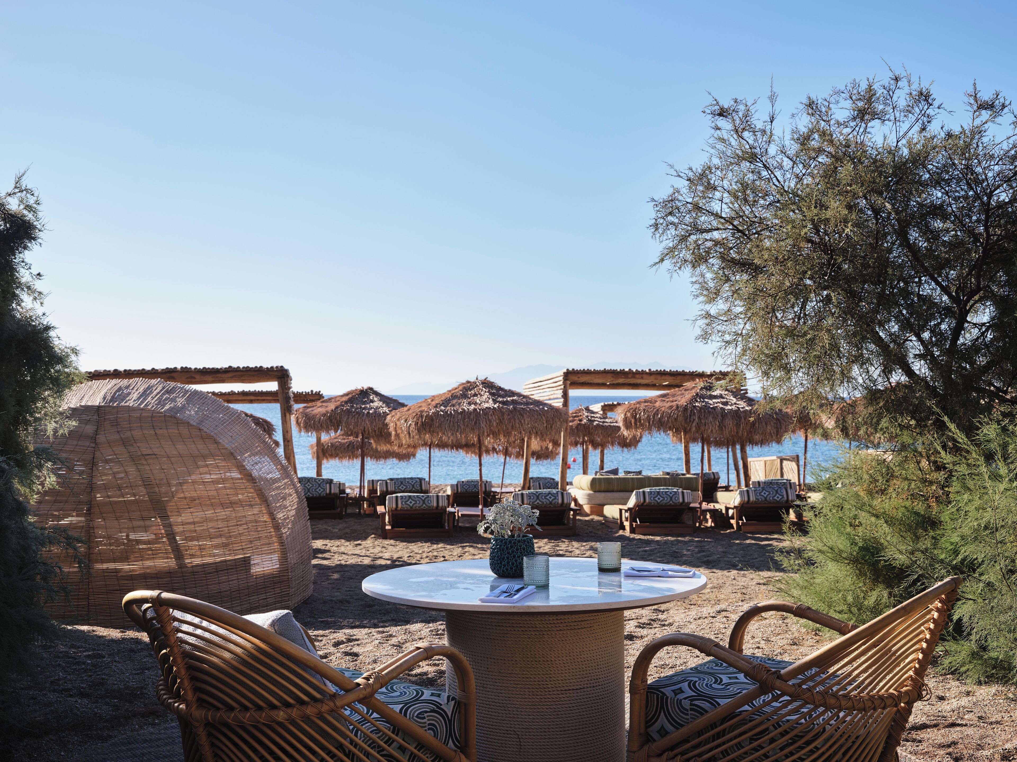 A beachside seating area with wicker furniture and thatched umbrellas, overlooking a calm sea.