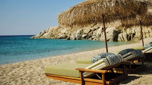 A beach with lounge chairs and a thatched umbrella. - Myconian Sunrise Relais & Chateaux Hotel (Mykonos)
