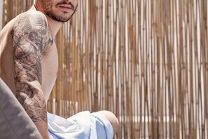 Pessoa com tatuagem no braço sentada em frente a uma cerca de bambu.