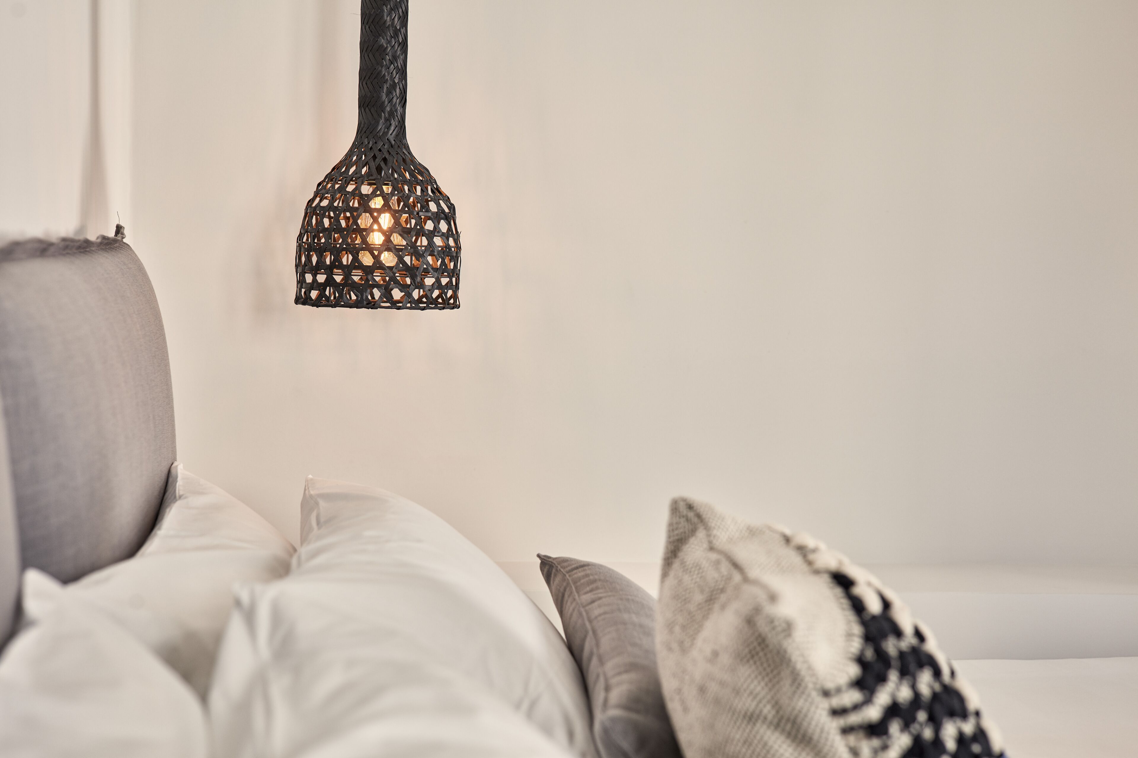 Una cama con ropa de cama blanca y gris, un cojín decorativo estampado y una lámpara colgante con diseño perforado.