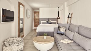 A modern bedroom with a bed, a grey sofa, a round coffee table, and a small round ottoman. - Boho Suites (Mykonos)