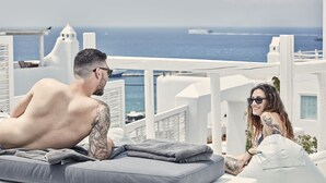A couple relaxing on a sun lounger with a view of the sea and white buildings. - Boho Suites (Mykonos)