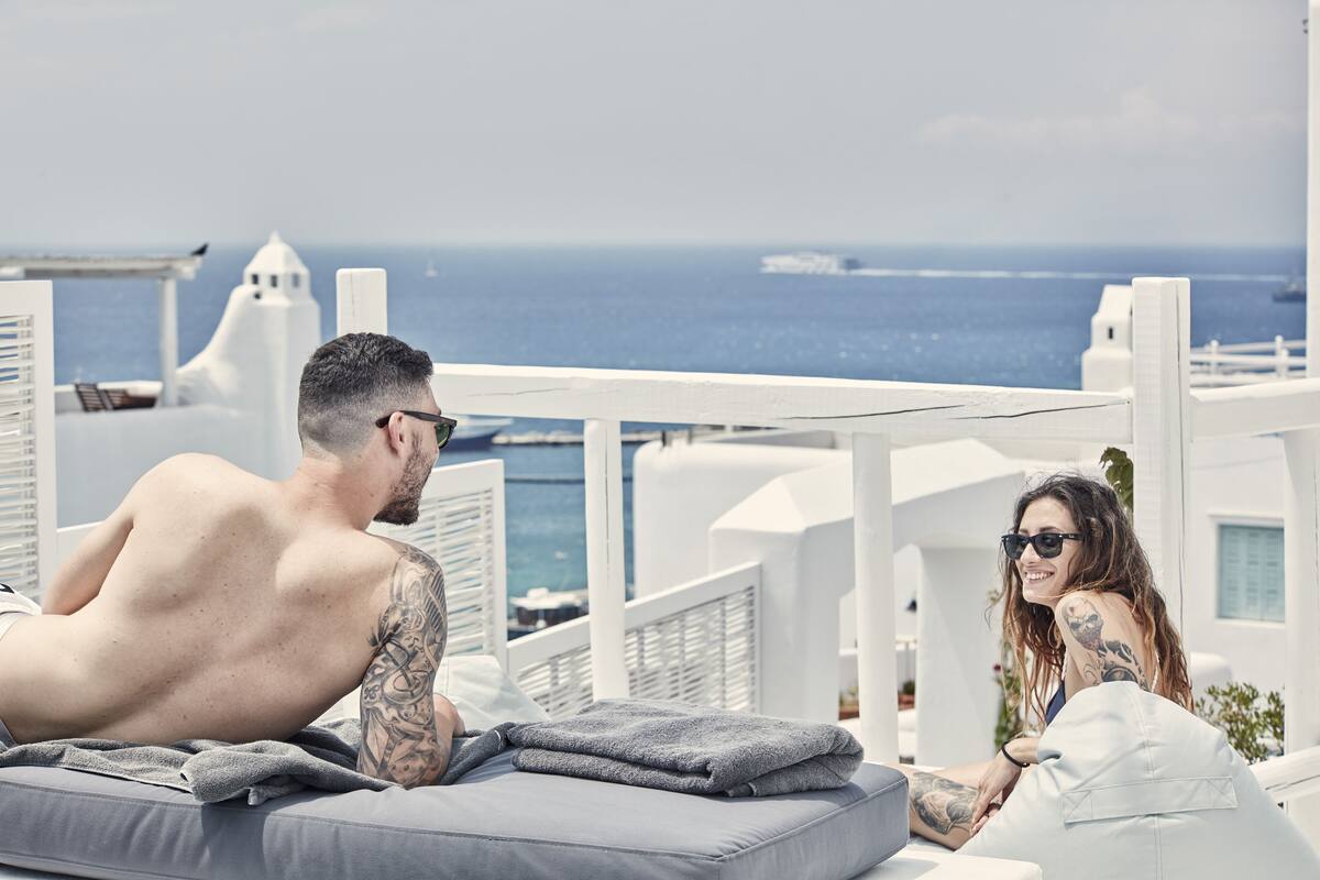 A couple relaxing on a sun lounger with a view of the sea and white buildings.