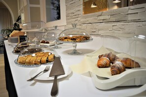 Una mesa de buffet con diversos pasteles y postres bajo campanas de cristal, una copa de vino y una cesta de cruasanes.