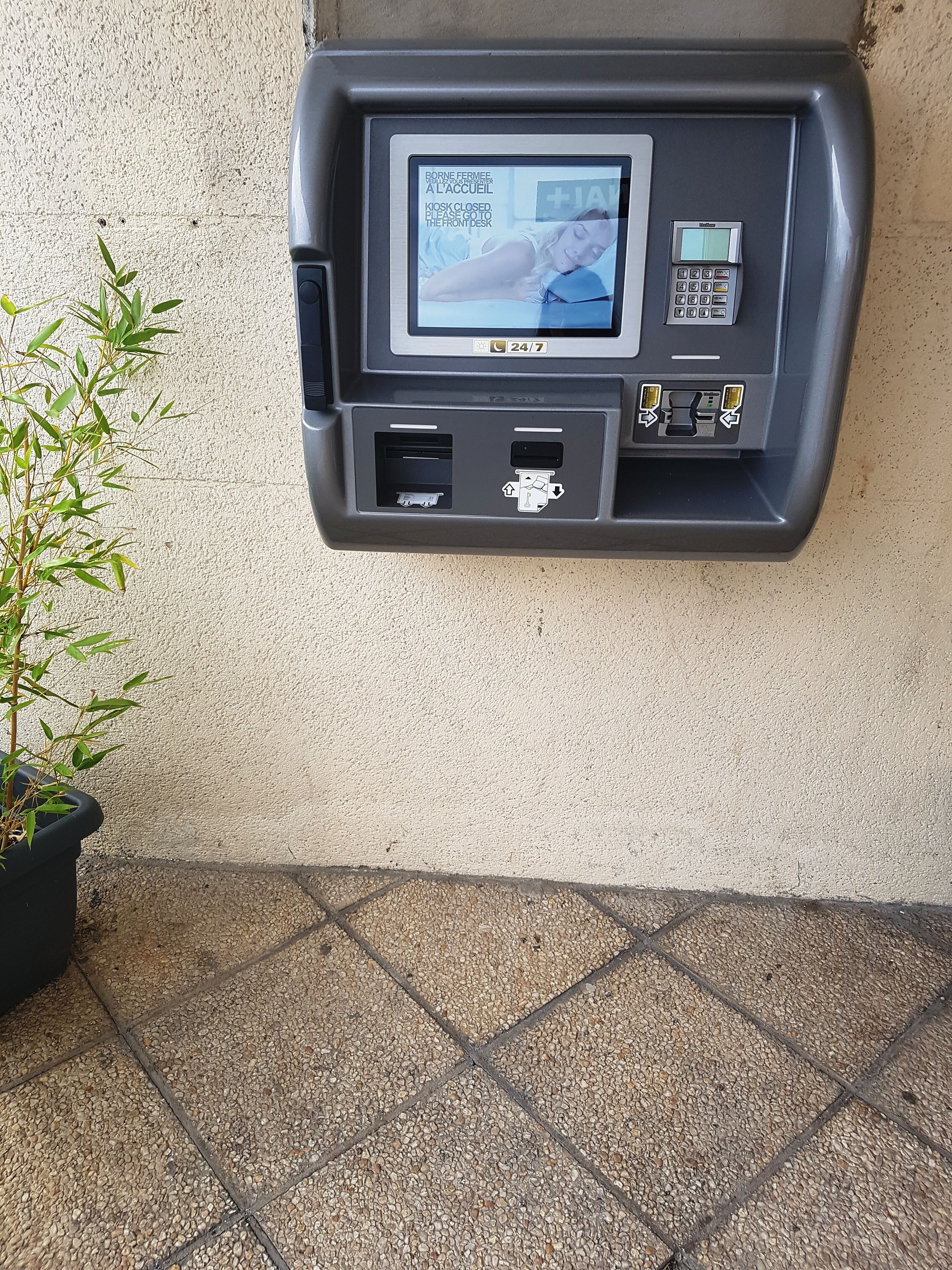 Un distributeur automatique de billets accessible 24 heures sur 24, 7 jours sur 7, fixé à un mur, avec un pot de plante placé à côté.
