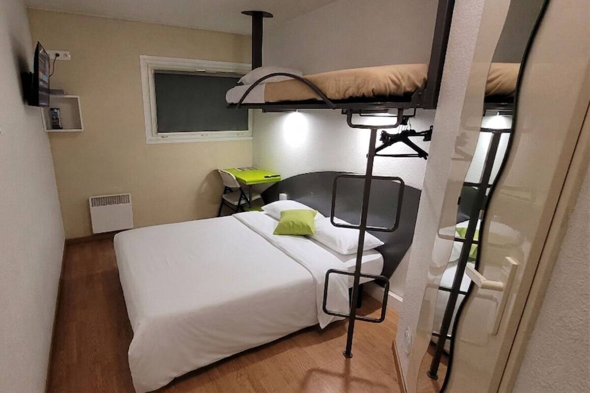 A compact hotel room with a bunk bed, a desk with a chair, a window, and a wall-mounted heater.