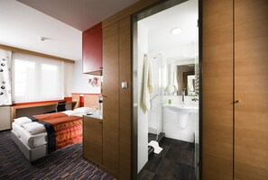 Habitación de hotel con cama, escritorio, silla, baño con lavamanos y espejo, y ducha.