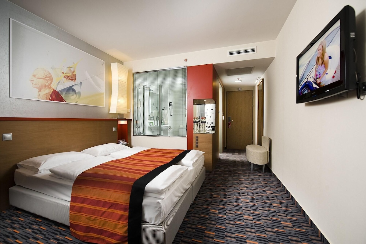 A hotel room with a bed, a television, a bathroom with a shower, and a small seating area.