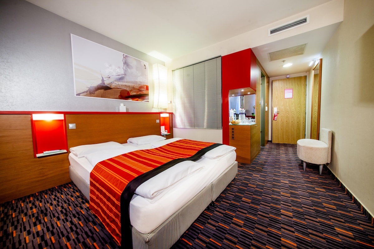 A hotel room with a large bed, a wooden headboard, a red accent wall, a patterned carpet, and a painting on the wall.