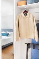 Una chaqueta color arena colgada de una percha en una habitación de hotel, con una cama, un almohadón azul y una maleta sobre una estantería.