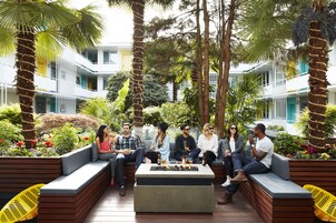 A group of people sitting on a wooden bench and a fire pit in a courtyard with plants and a building in the background.