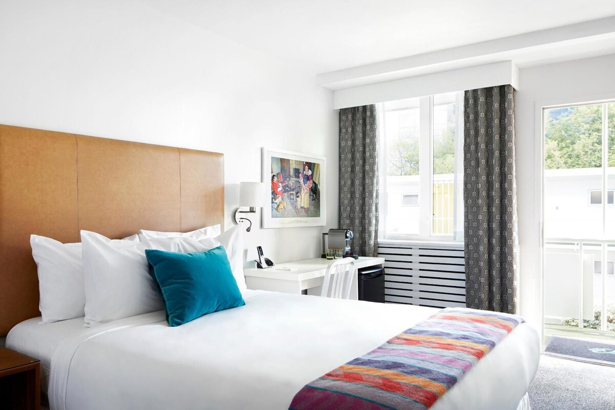 A hotel room with a bed, a desk, a chair, a window with curtains, and a picture on the wall.