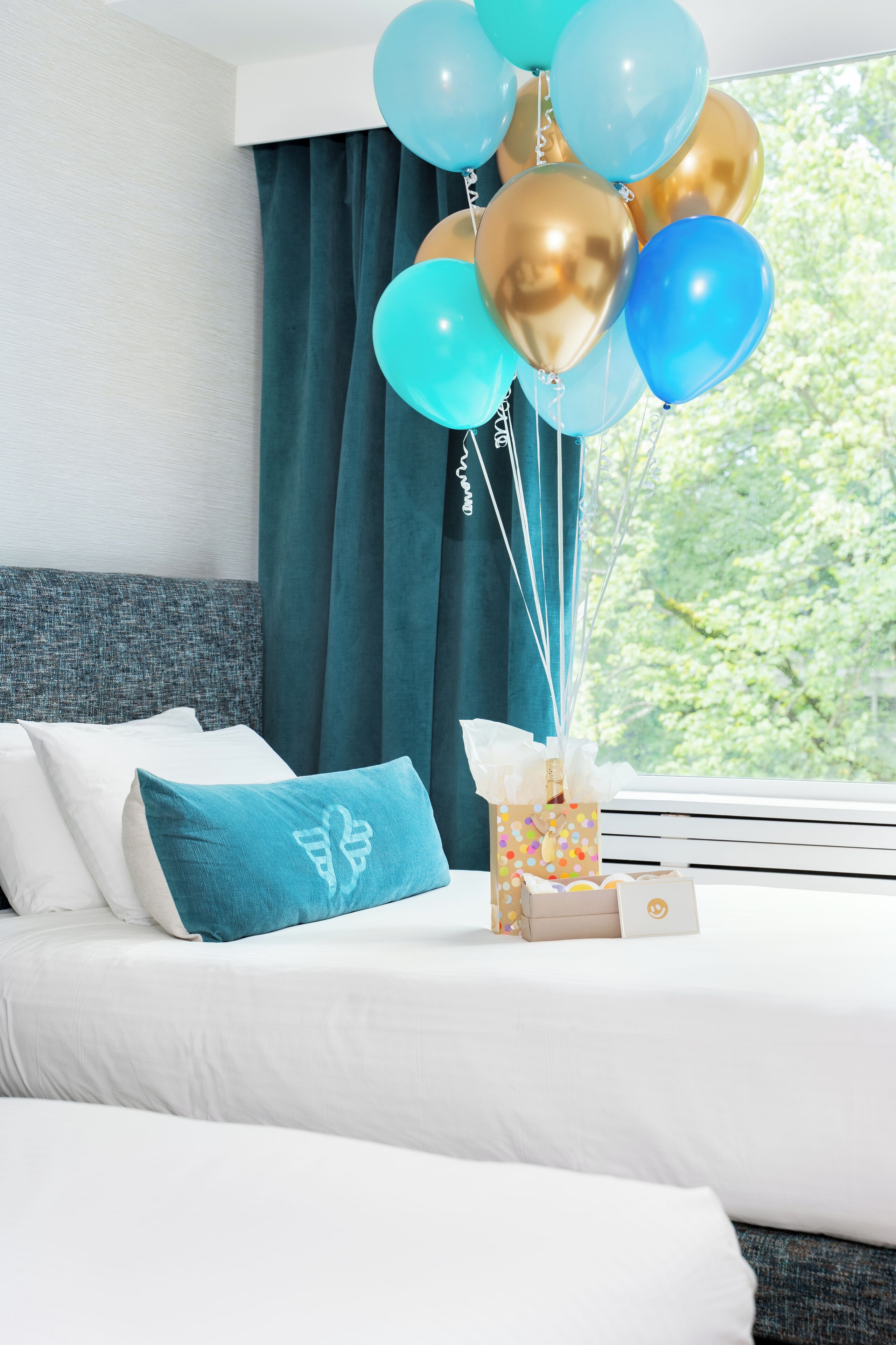 A hotel room with a bed, teal curtains, and a window with a view of trees.