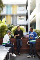Three men standing and sitting on a bench in an outdoor area with a modern building in the background.