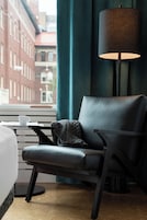 Habitación de hotel con cama, una silla, una mesa, una lámpara y vistas de edificios a través de una ventana.