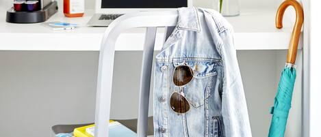 Un escritorio blanco con una computadora portátil, una cafetera, una botella, una planta, una chaqueta vaquera y un paraguas.