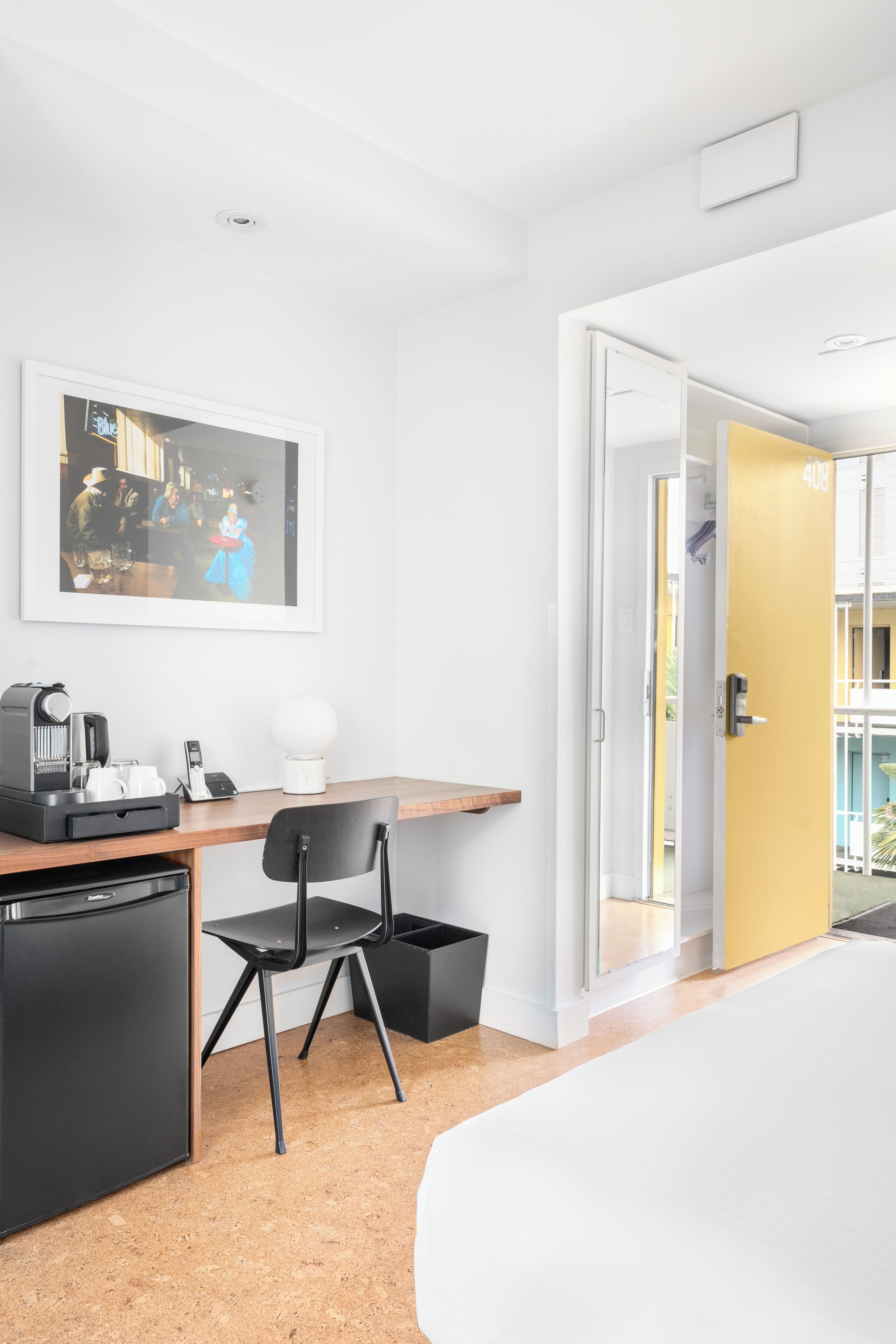 Une chambre d’hôtel avec un lit, un bureau doté d’une machine à café, une chaise, une poubelle et un tableau encadré au mur.
