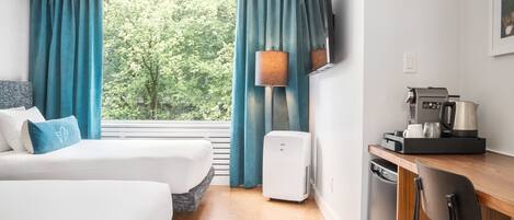 A hotel room with two beds, a desk with a coffee maker, a TV, and a view of trees through the window.
