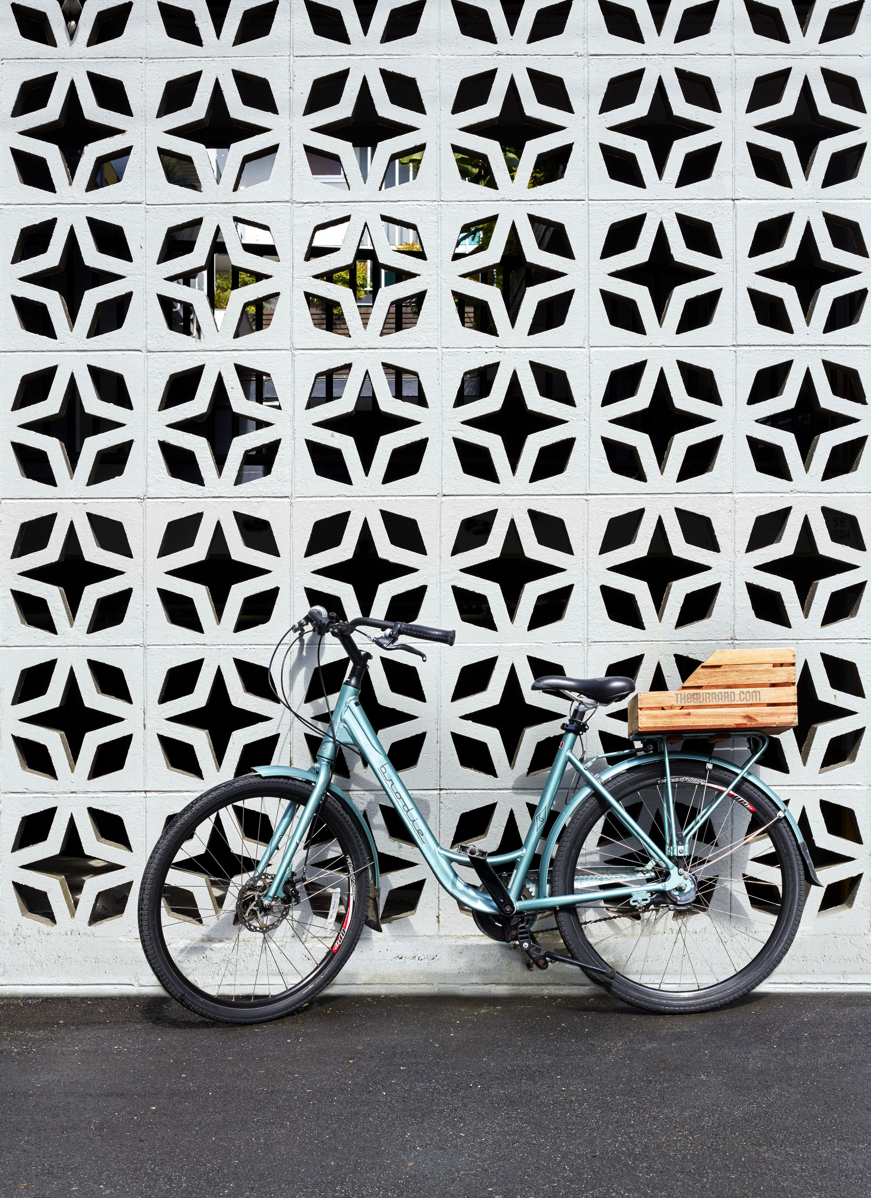 Een smaragdgroene fiets met een houten doos bevestigd aan de achterkant, geparkeerd tegen een witte muur met een geometrisch patroon.