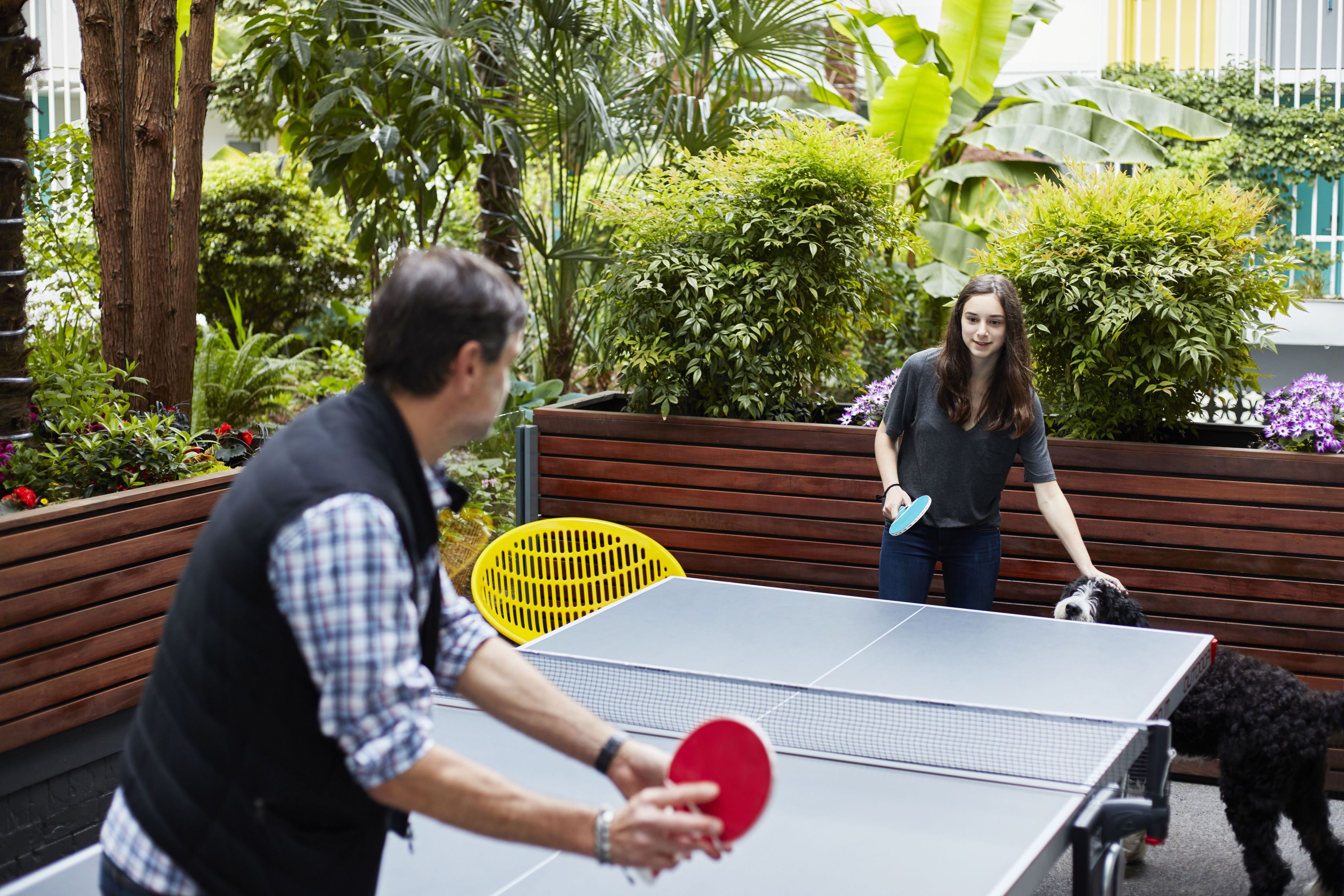 Mesa de ping pong en un espacio exterior con bancos de madera y follaje.