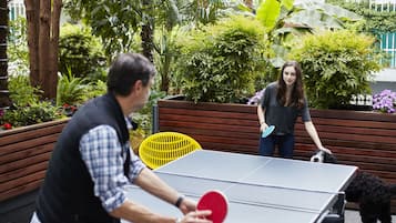 Mesa de ping pong en un espacio exterior con bancos de madera y follaje.