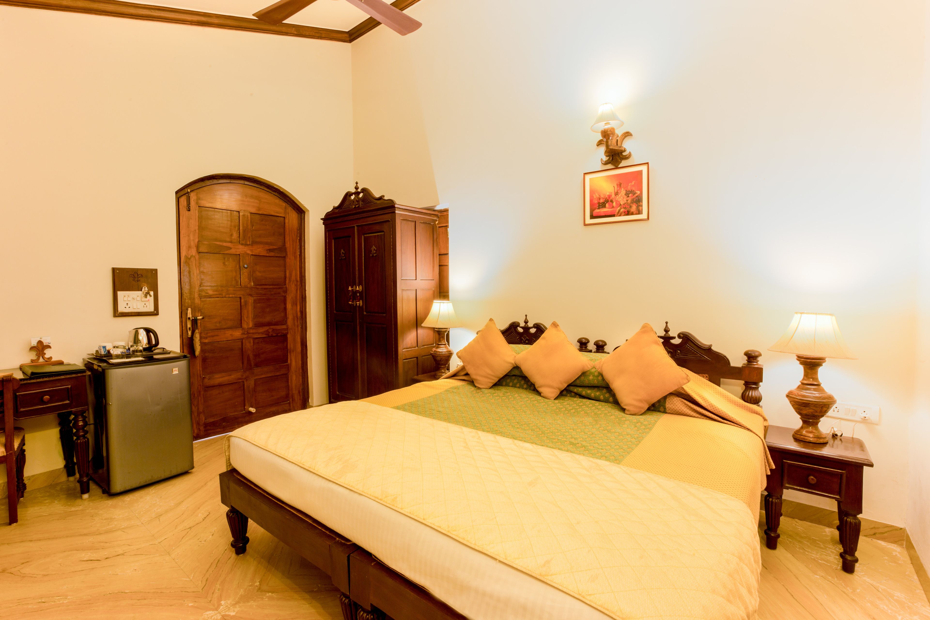 A neatly arranged bedroom with a large bed, a wooden wardrobe, a desk with a lamp, and a small refrigerator.