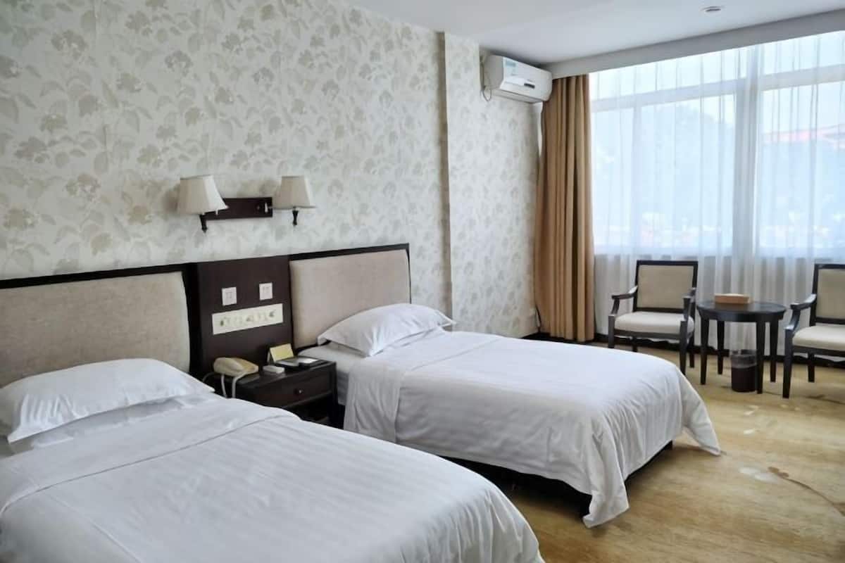 A hotel room with two beds, a nightstand, a desk with chairs, and a large window with curtains.