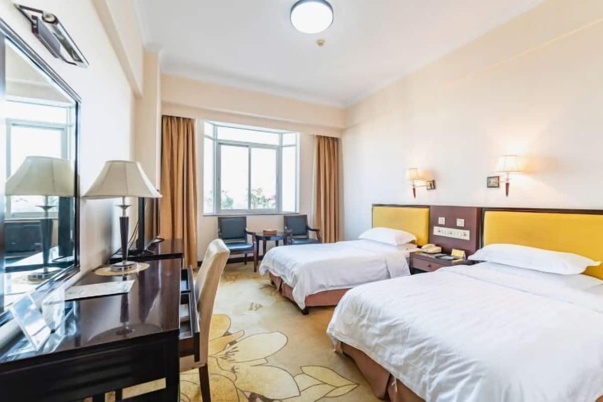 A hotel room with two beds, a desk with a lamp, a chair, a small table, and a window with curtains.