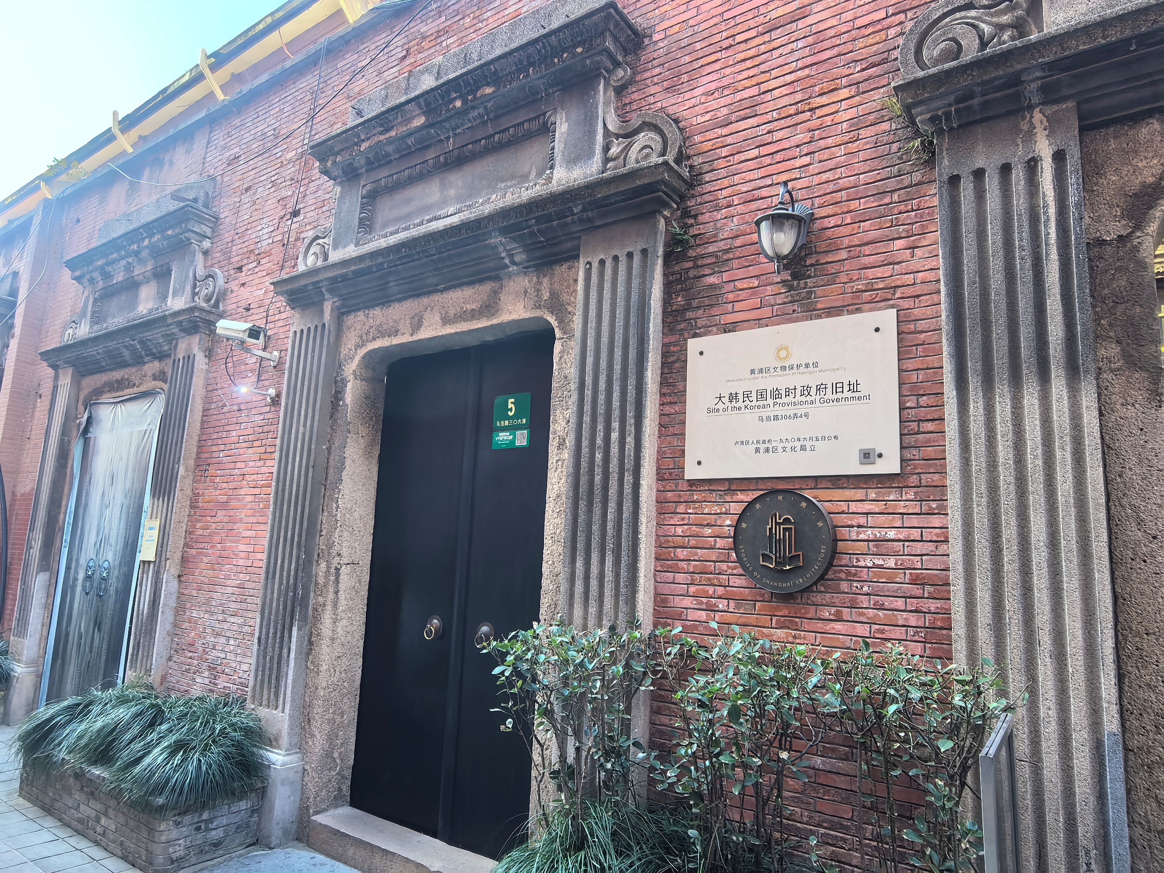 Un edificio histórico con una puerta negra, una placa que lo identifica como sede del Gobierno Provisional Coreano, y un pequeño jardín con plantas.