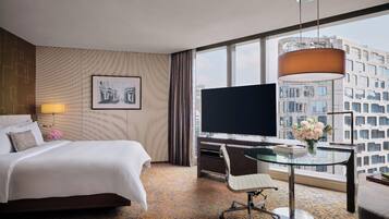 A hotel room with a bed, a desk with a chair, a TV, a large window, and a city view.