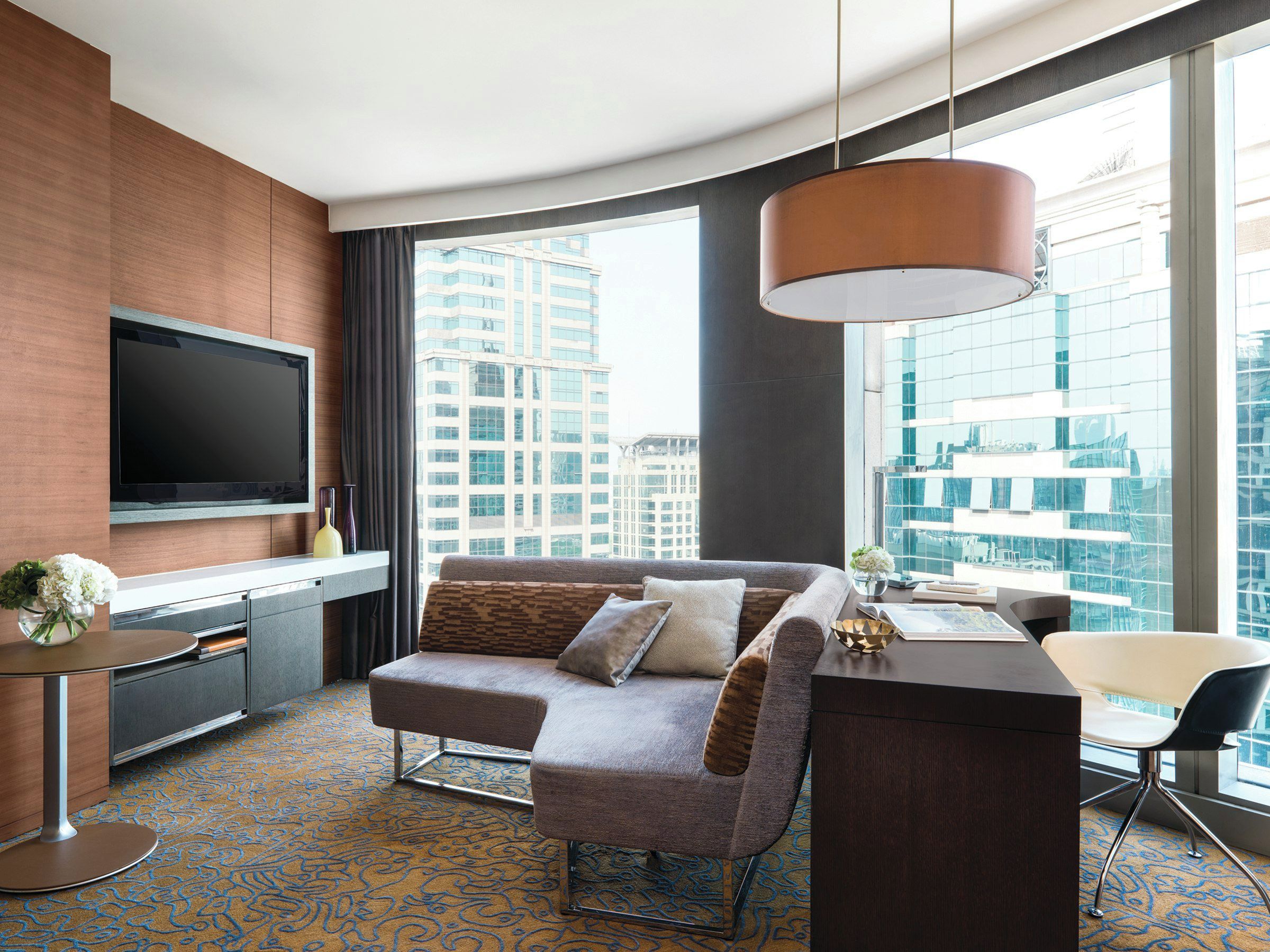 A modern hotel room with a large window, a flat-screen TV, a sofa, a desk, and a chair.