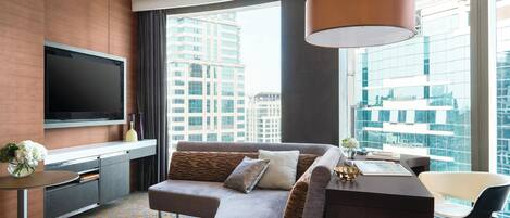 A modern hotel room with a large window, a flat-screen TV, a sofa, a desk, and a chair.