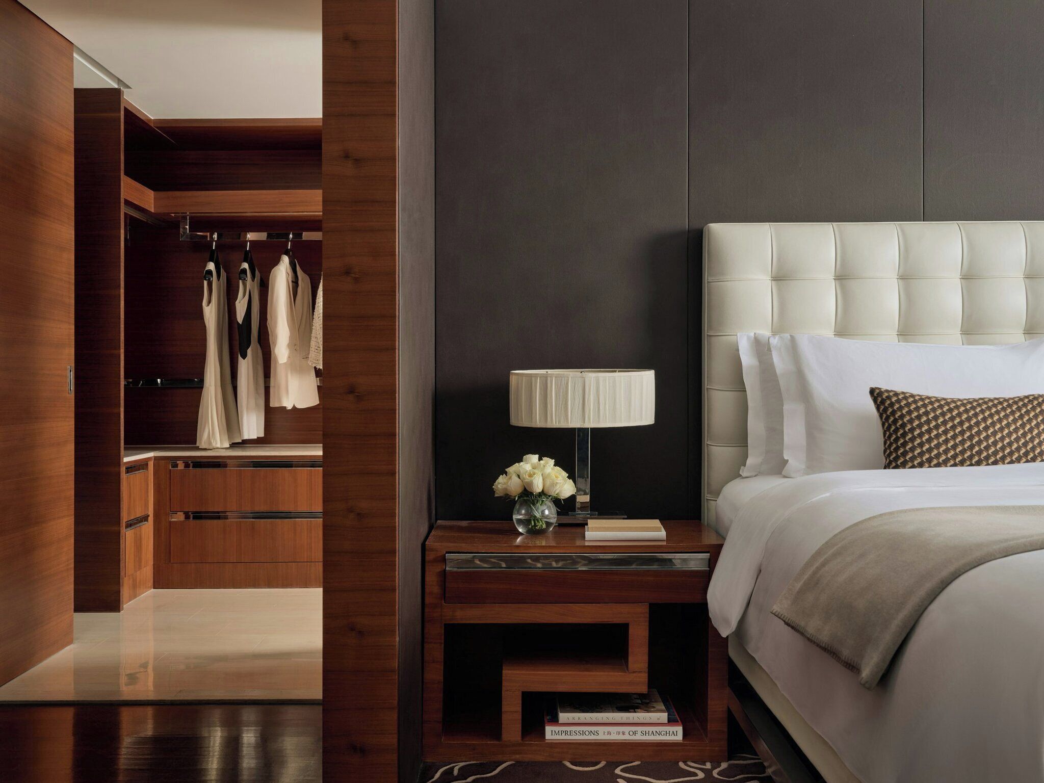 A modern hotel room with a wooden wardrobe, a bedside table with a lamp and books, and a large bed with a tufted headboard.
